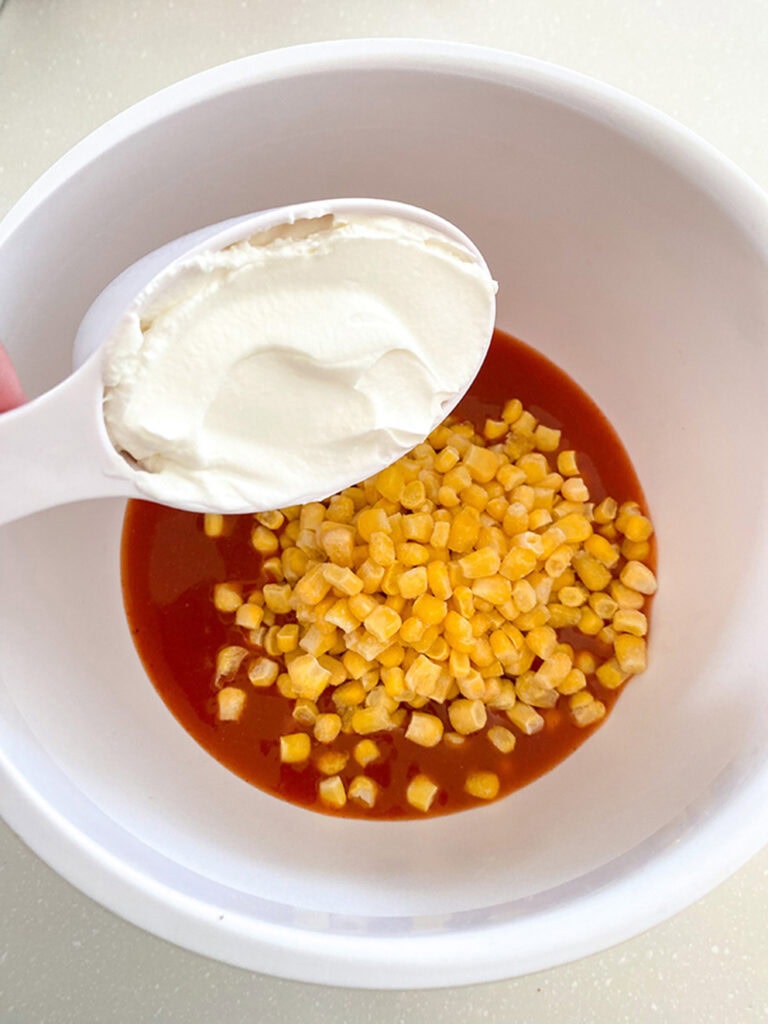a white bowl of enchilada sauce, corn and sour cream being added