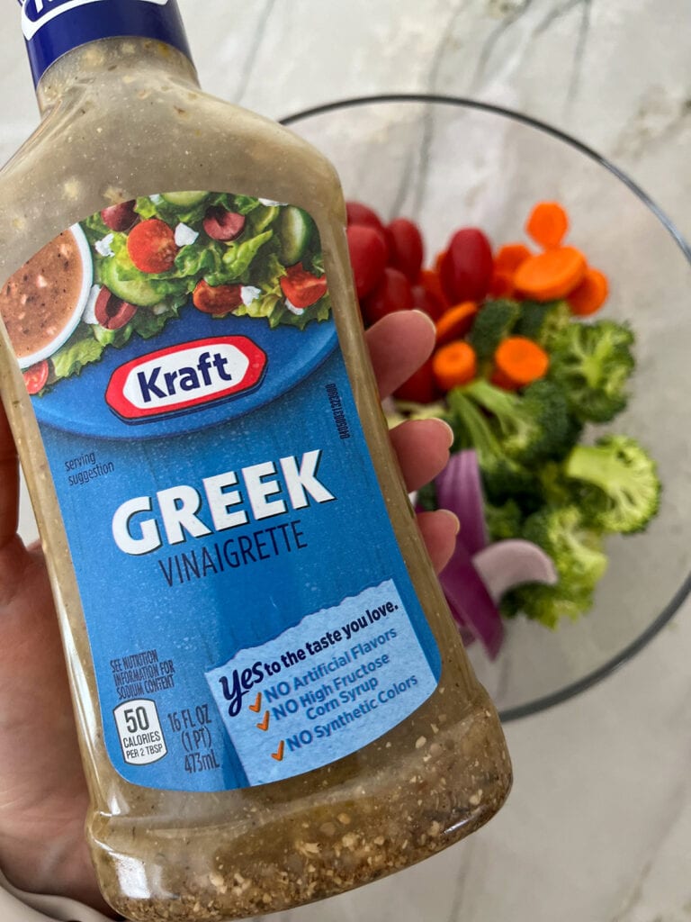 bottle of Greek salad dressing with a bowl of veggies in distance
