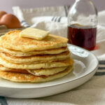 a stack of Homemade Pancakes with butter on a white plate with syrup, and eggs in background on a blue and white towel