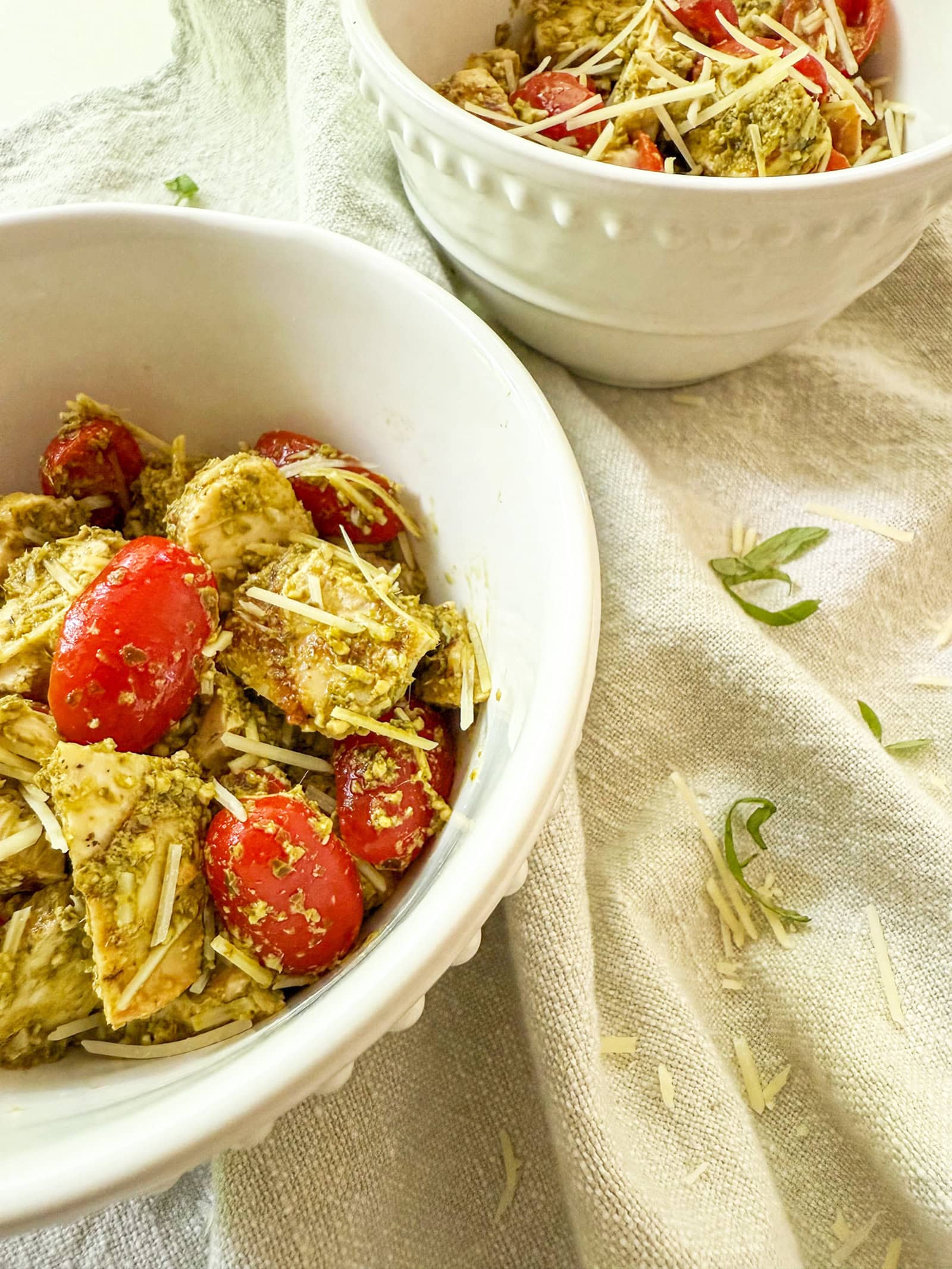 two servings of Pesto Chicken Salad recipe in textured white bowls on a neutral colored tablecloths. Extra fresh basil is sprinkled as garnish
