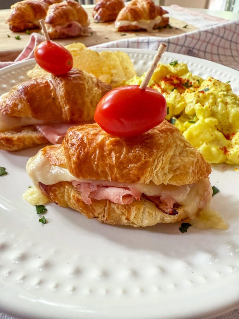 Mini Ham and Cheese Croissant Sandwiches on a white plate with a tomato garnish and side of potato salad and chips