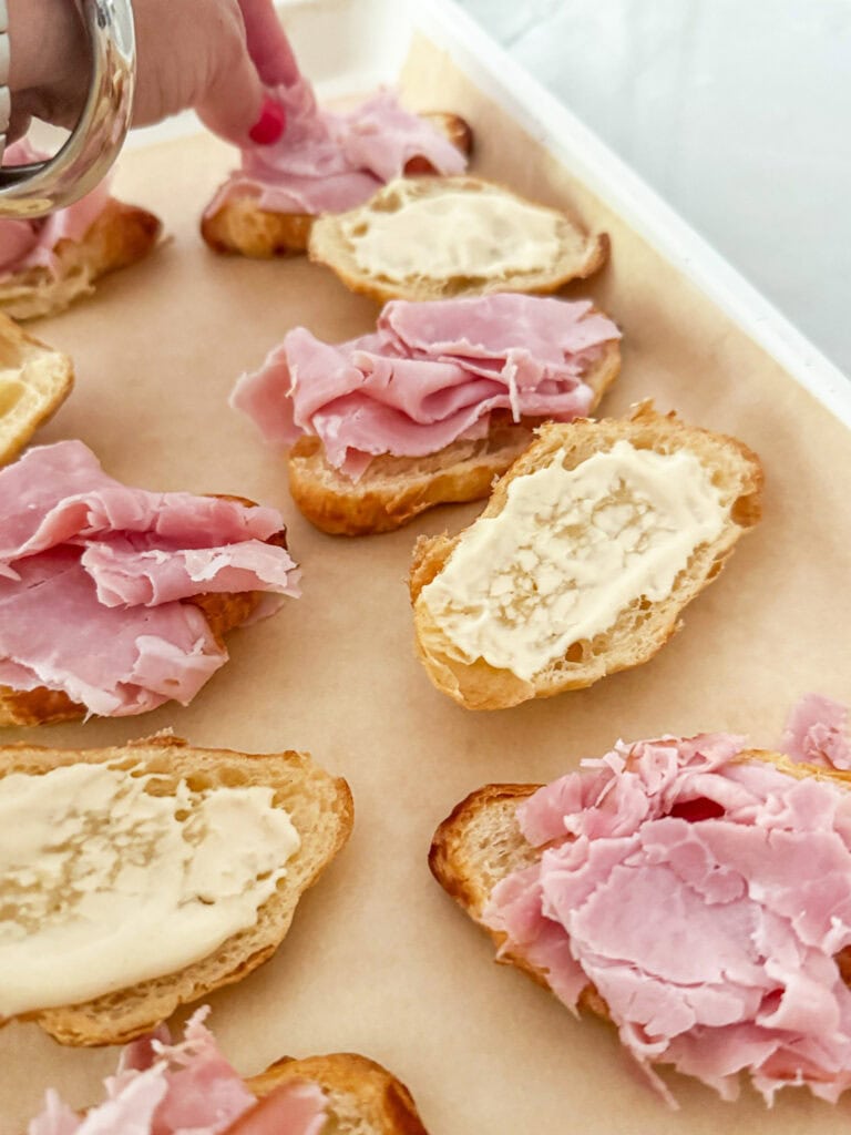 sliced croissants on a parchment paper lined baking sheet with mayo-mustard spread one one side and ham on the other