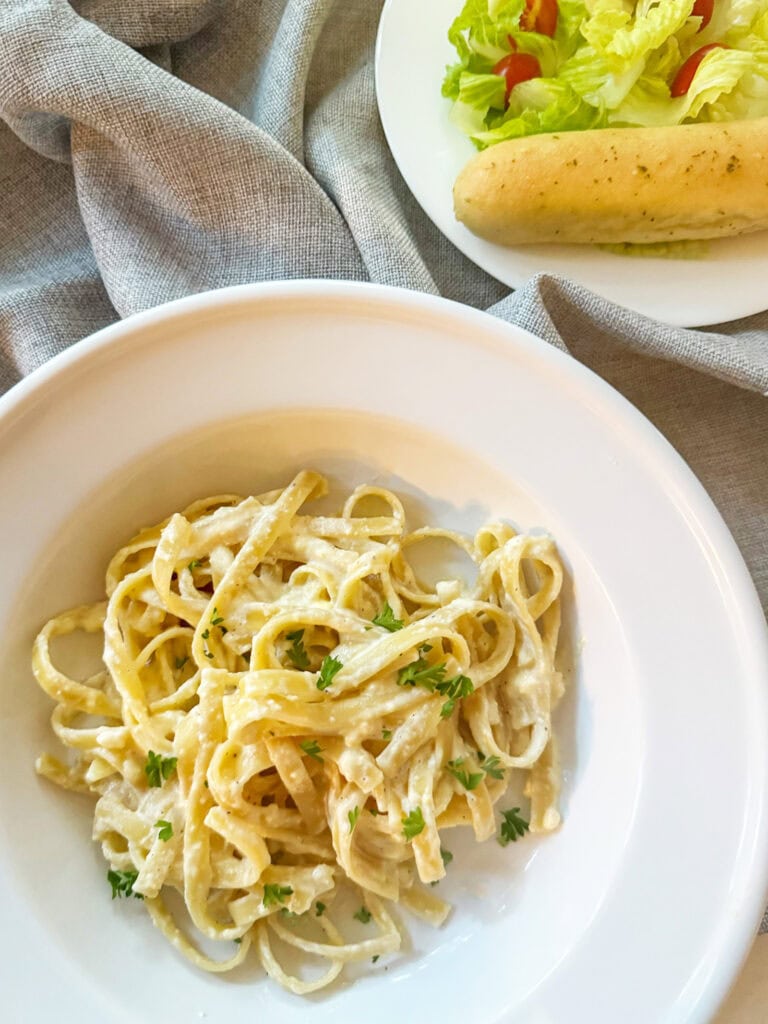 Copy Cat Olive Garden Fettuccine Alfredo in a white bowl with a salad and breadstick in the background.