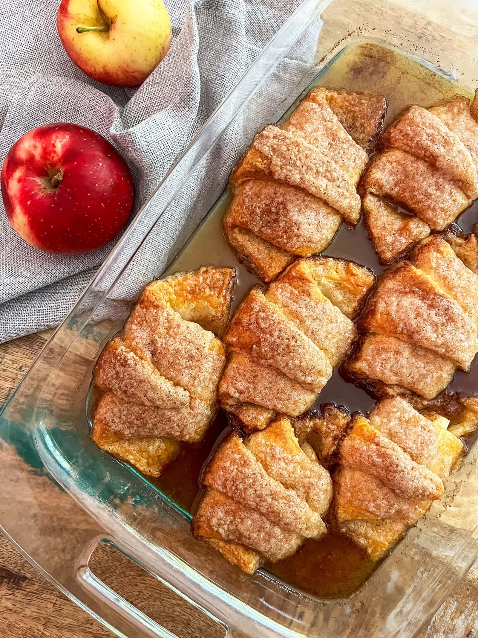 baked Crescent Roll Apple Dumplings in glass pan on wood table with red apple and grey napkin