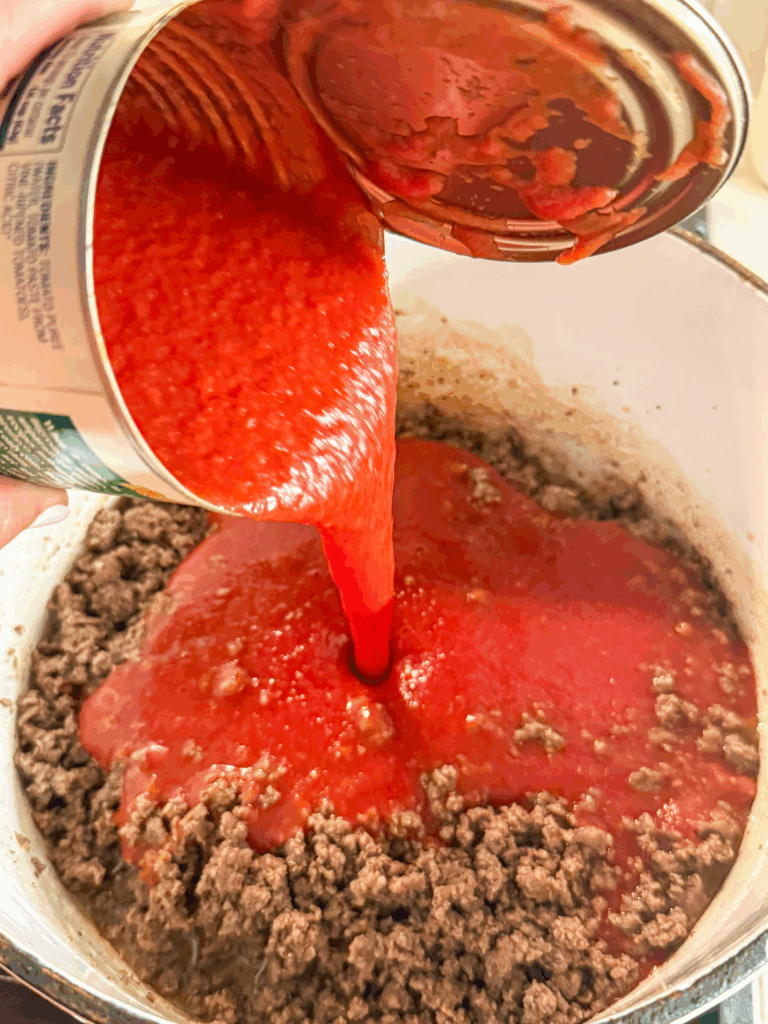pouring tomato sauce into pot of browned grown beef