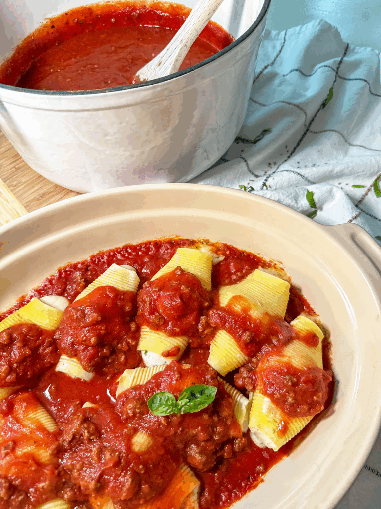 Homemade Italian Meat Sauce served over cheese stuffed pasta shells in a baking dish with basil garnish and extra sauce in white Dutch oven in background