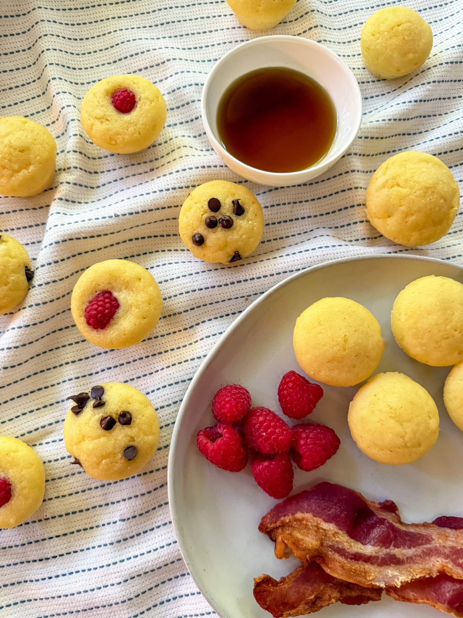 mini pancake bites on a plate with bacon and berries and a side of maple syrup with extra pancake bites sprinkled around a white and blue napkin