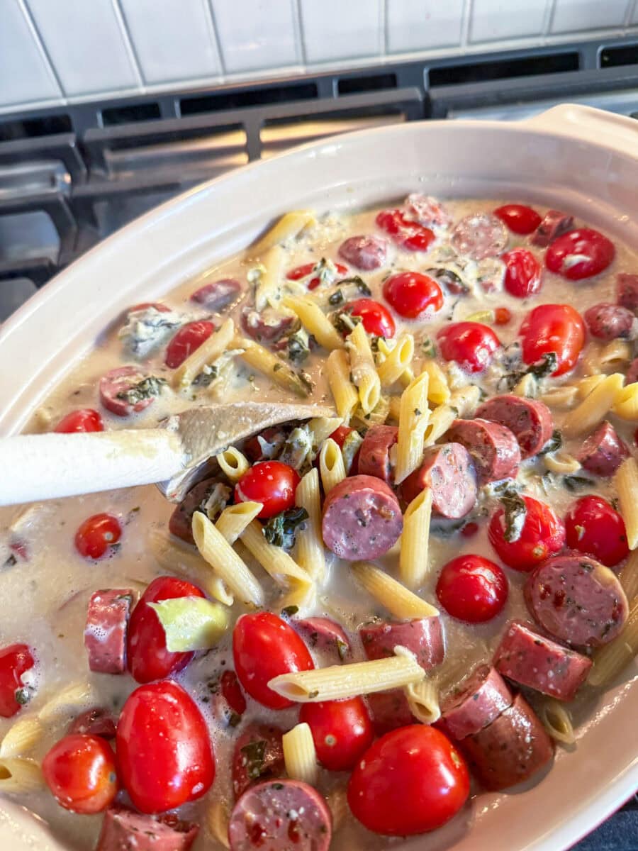 a wooden spoon stirring Spinach and Artichoke Dip Pasta Bake with Chicken Sausage