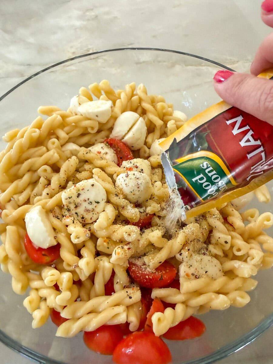 adding italian dressing seasoning to Caprese Pasta Salad in a glass bowl