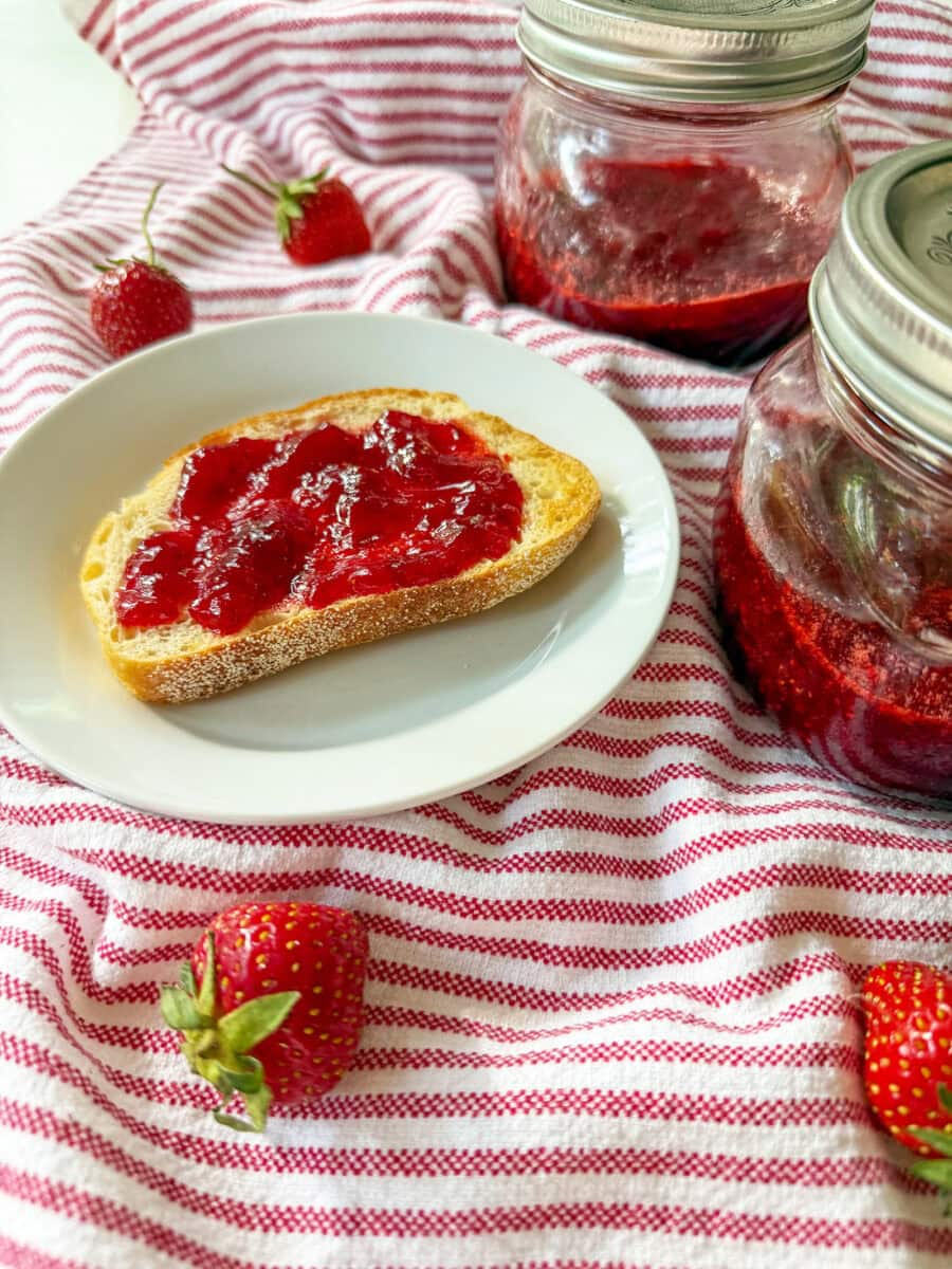 Homemade Strawberry Jam (No Pectin) on toast with mason jars of jam on red and white striped napkin with fresh strawberries sprinkled around