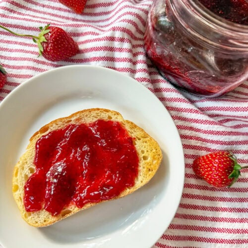 Homemade Strawberry Jam (No Pectin) on sourdough toast with mason jars of jam on red and white striped napkin with fresh strawberries sprinkled around