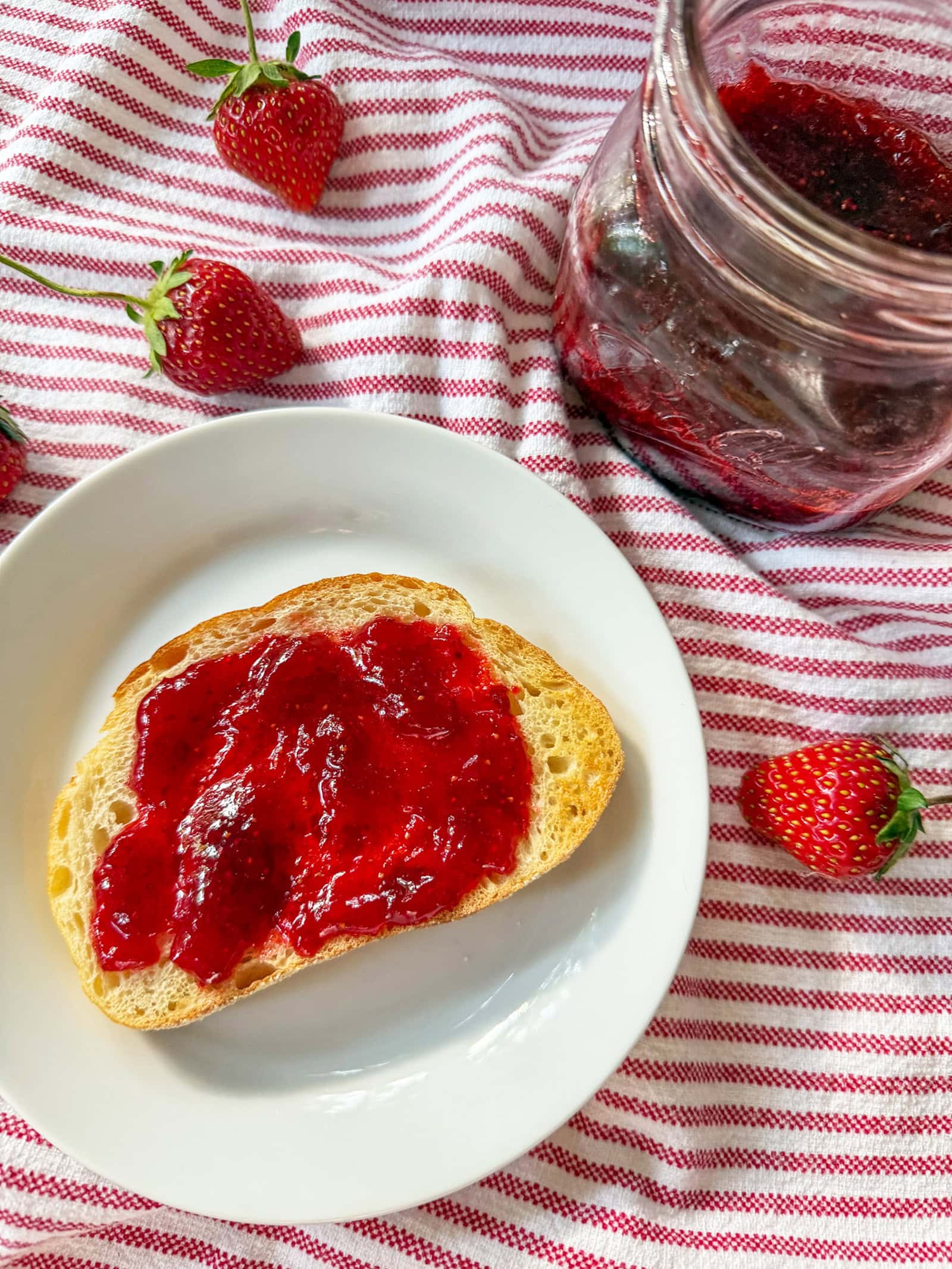Homemade Strawberry Jam (No Pectin) on sourdough toast with mason jars of jam on red and white striped napkin with fresh strawberries sprinkled around